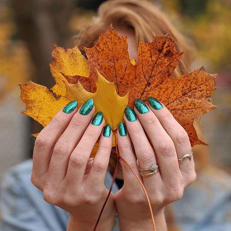 Person holding autumn leaves with sparkling teal nail art against a blurred outdoor background.