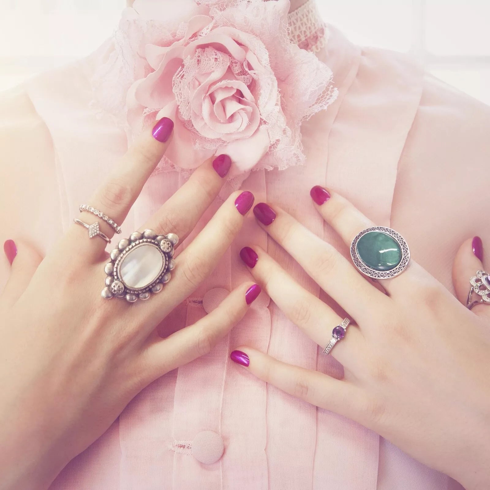 Elegant hands adorned with rings, showcasing vibrant pink manicure against a soft pink blouse.